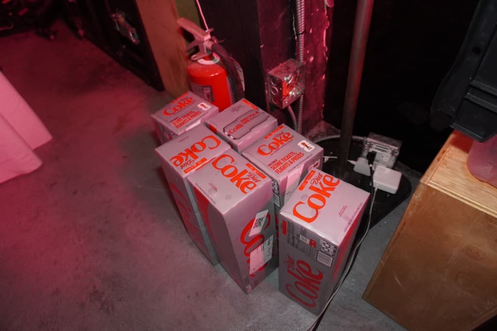Stacked Diet Coke cartons on concrete, red backstage wash.
